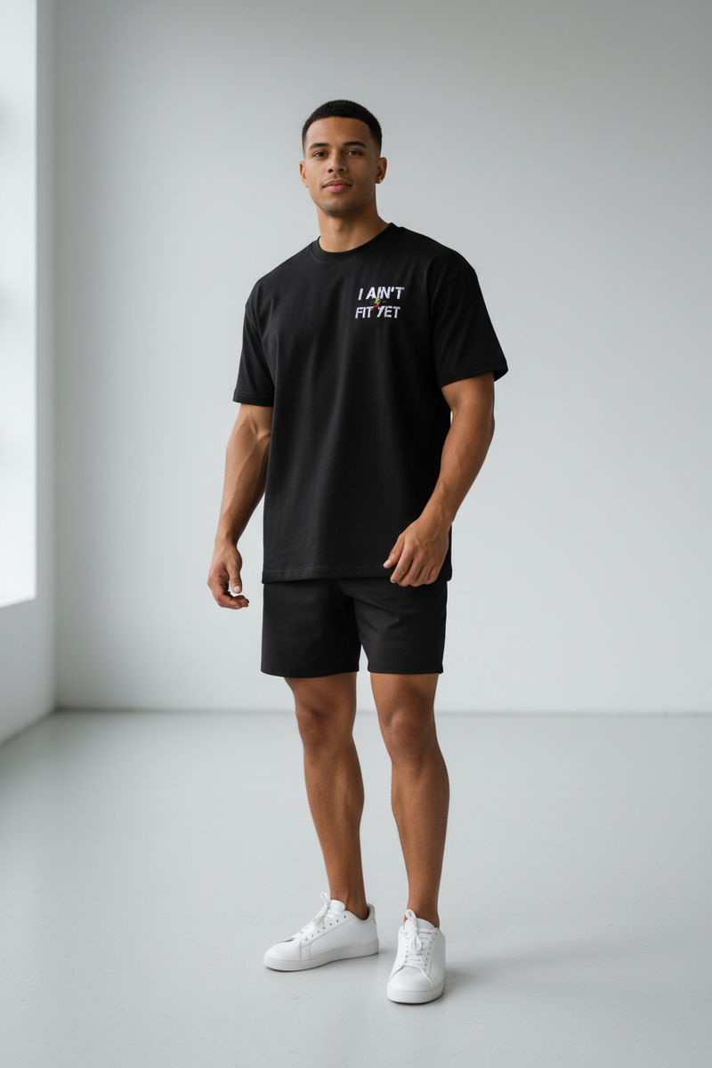 A man modeling a black t-shirt with the text 'I AM NOT A FIT' in white on the chest, paired with black athletic shorts and white sneakers, standing in a minimalist studio setting.