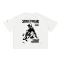 An off-white short-sleeve t-shirt with a black graphic print on the front. The graphic features the text 'STREETWEAR' and 'URBAN FASHION' surrounding a stylized illustration of a person sitting on the ground with a skateboard.