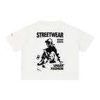 An off-white short-sleeve t-shirt with a black graphic print on the front. The graphic features the text 'STREETWEAR' and 'URBAN FASHION' surrounding a stylized illustration of a person sitting on the ground with a skateboard.