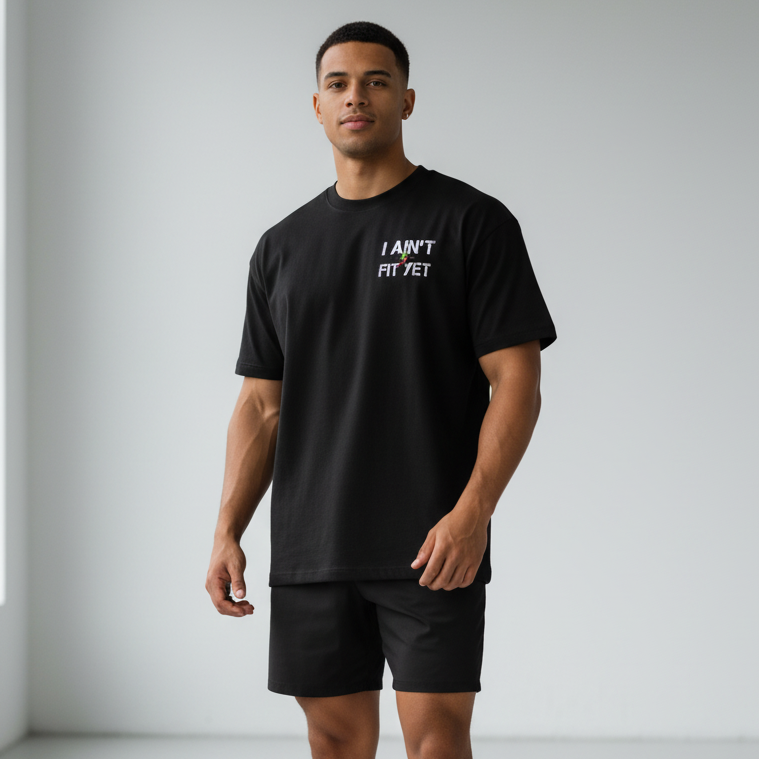 A man modeling a black t-shirt with the text 'I AM NOT A FIT' in white on the chest, paired with black athletic shorts and white sneakers, standing in a minimalist studio setting.