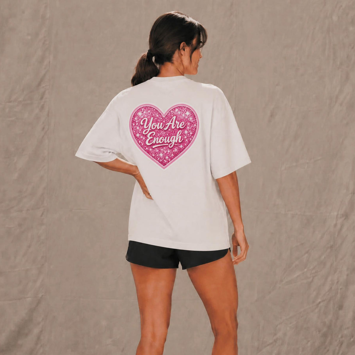 A woman shown from the back wearing an oversized white T-shirt with a large pink glittery heart graphic on the back that reads 'You Are Enough' in white script. She is also wearing black shorts, standing against a textured gray backdrop.