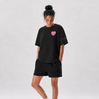 A model wearing a black oversized t-shirt with a pink heart graphic on the chest, matching black shorts, white socks, and white New Balance sneakers, standing against a light gray studio background.